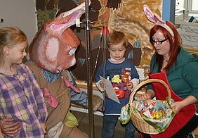 Ein Kind steht mit einem Blatt Papier in der Mitte, während ein Mensch mit einem Hasenkostüm und zwei Frauen mit Hasenohren um ihn herum stehen. Eine der Frauen hält einen Korb mit bunten Eiern. Die Szene vermittelt eine festliche und fröhliche Atmosphäre.