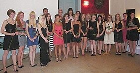 Eine Gruppe von jungen Frauen steht zusammen in eleganter Abendkleidung. Viele halten eine Rose in der Hand. Der Hintergrund ist ein festlich dekorierter Raum, und alle Personen scheinen feierlich und erfreut zu sein.