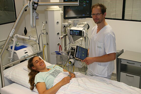 In einem Krankenhauszimmer liegt eine lächelnde Patientin im Bett. Daneben steht medizinisches Personal in weißer Kleidung und überwacht mit Tablet und Monitoren die Vitalwerte. Die angeschlossenen Geräte deuten auf eine laufende Untersuchung oder Behandlung.