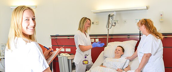 Im Krankenhauszimmer betreuen drei Pflegekräfte eine lächelnde Patientin im Bett. Eine schreibt Notizen, eine trägt Handschuhe, eine spricht beruhigend. Die Atmosphäre wirkt freundlich und professionell.