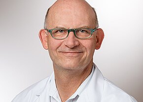 Porträt eines älteren Mannes mit Glatze und türkisfarbener Brille, der freundlich lächelt und einen weißen Kittel trägt; neutraler Hintergrund.