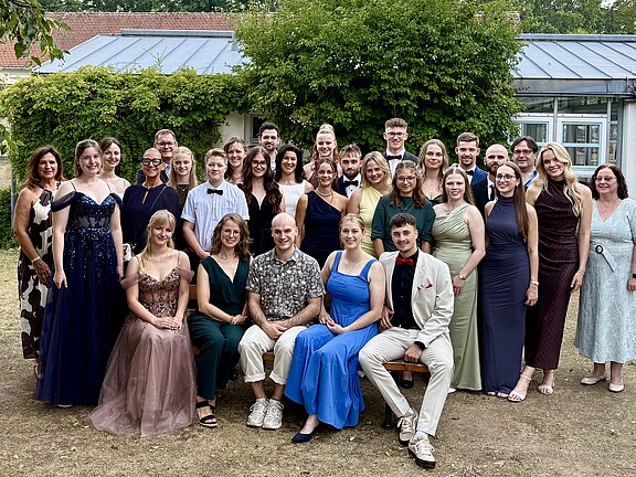 Gruppenfoto von rund 30 festlich gekleideten Erwachsenen, die draußen vor einem Gebäude in mehreren Reihen posieren und lächeln. Einige tragen Abendkleider, andere Anzüge; zwei sitzen vorn auf einer Bank. Sommerliche Atmosphäre mit Bäumen und Efeu im Hintergrund.