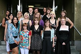 Eine Gruppe von 15 Personen steht lächelnd auf einer Treppe vor einem Gebäude. Die Frauen tragen verschiedene Outfits, von casual bis elegant. Es herrscht eine fröhliche Stimmung, und alle scheinen sich zu amüsieren.