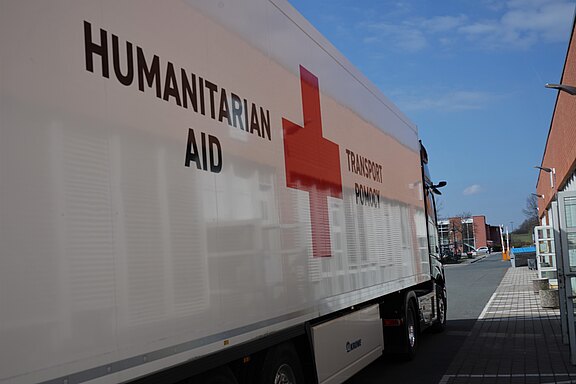 Lkw mit großem Rotkreuz‑Logo und der Aufschrift „Humanitarian Aid“. Er steht auf einer Straße und dient dem Transport von Hilfsgütern.