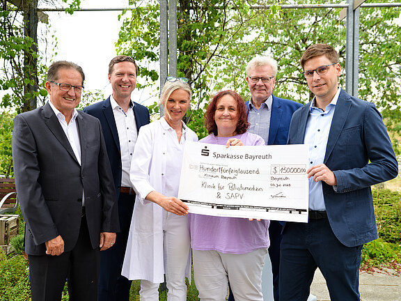 Eine Gruppe von fünf Personen steht im Freien und hält einen großen Scheck der Sparkasse Bayreuth über 150.000 Euro. Der Scheck ist für die Klinik für Palliativmedizin und SAPV bestimmt. Das Datum auf dem Scheck ist der 3. Juli 2023.