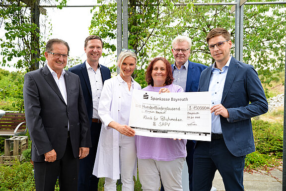 Eine Gruppe von fünf Personen steht im Freien und hält einen großen Scheck der Sparkasse Bayreuth über 150.000 Euro. Der Scheck ist für die Klinik für Palliativmedizin und SAPV bestimmt. Das Datum auf dem Scheck ist der 3. Juli 2023.