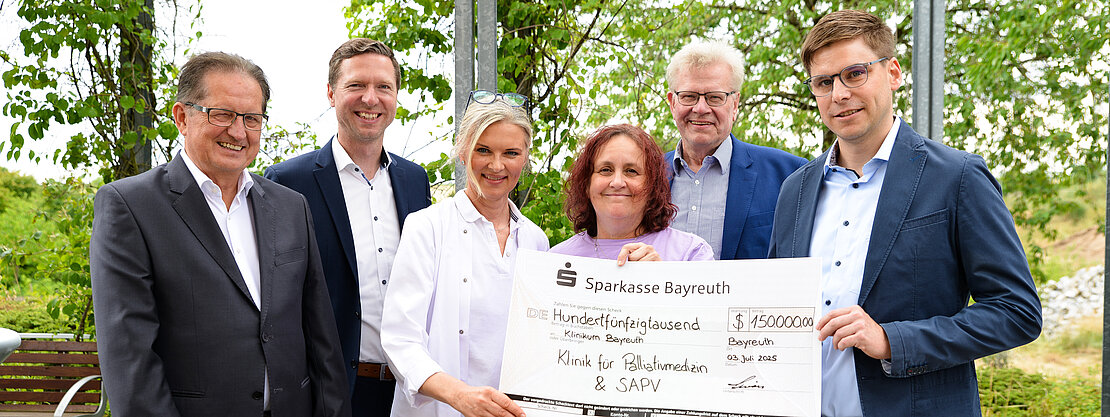 Eine Gruppe von fünf Personen steht im Freien und hält einen großen Scheck der Sparkasse Bayreuth über 150.000 Euro. Der Scheck ist für die Klinik für Palliativmedizin und SAPV bestimmt. Das Datum auf dem Scheck ist der 3. Juli 2023.