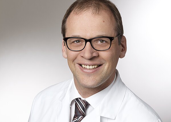 Porträt eines lächelnden Mannes mit dunkler Brille und kurzem Haar. Er trägt weißen Arztkittel, weißes Hemd und gestreifte Krawatte. Neutraler, hellgrauer Hintergrund.