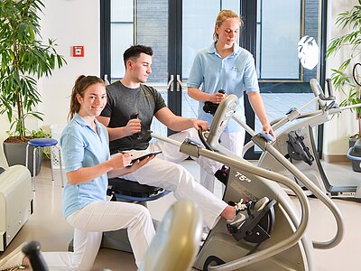 Zwei Physiotherapeutinnen betreuen einen Mann beim Training auf einem Fitnessgerät. Die eine Therapeutin notiert etwas, während die andere ihm hilft. Der Raum ist hell und freundlich gestaltet, mit Pflanzen und großen Fenstern.