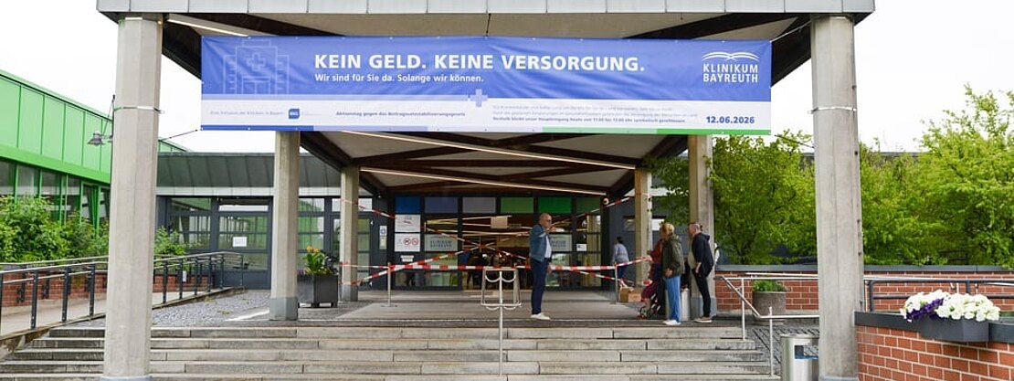 Eingang eines Krankenhauses mit großem blauen Banner: Kein Geld. Keine Versorgung. Aufschrift nennt Klinikum Bayreuth und ein Datum. Einige Personen stehen vor dem Eingang. Das Transparent warnt vor fehlender Finanzierung und eingeschränkter Versorgung.