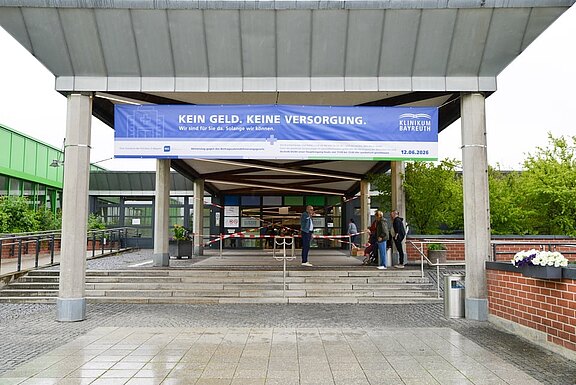 Eingang eines Krankenhauses mit großem blauen Banner: Kein Geld. Keine Versorgung. Aufschrift nennt Klinikum Bayreuth und ein Datum. Einige Personen stehen vor dem Eingang. Das Transparent warnt vor fehlender Finanzierung und eingeschränkter Versorgung.