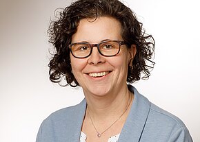 Melanie Häfner Porträtfoto einer lächelnden Person mit lockigen dunklen Haaren und Brille vor neutralem Hintergrund. Sie trägt eine hellblaue Jacke über einer weißen Bluse sowie eine Halskette mit kleinem Anhänger. Fokus auf freundliches, professionelles Auftreten.