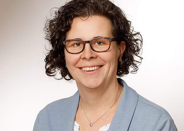 Porträtfoto einer lächelnden Person mit lockigen dunklen Haaren und Brille vor neutralem Hintergrund. Sie trägt eine hellblaue Jacke über einer weißen Bluse sowie eine Halskette mit kleinem Anhänger. Fokus auf freundliches, professionelles Auftreten.