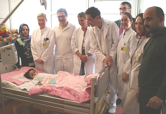 Ein kleines Kind liegt in einem Krankenhausbett mit rosa Decke. Mehrere Ärztinnen und Ärzte in weißen Kitteln stehen lächelnd daneben. Zwei Angehörige stehen am Bett. Die Szene wirkt wie ein Besuch oder eine Visite im Patientenzimmer.