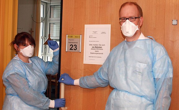 Zwei medizinische Fachkräfte in Schutzkleidung stehen an einer Krankenzimmertür. Sie tragen Kittel, Handschuhe und FFP-Masken. Eine weist auf ein Schild am Eingang hin, das vor Betreten um Anmeldung beim Stationspersonal bittet. Tür mit Zimmernummer 23.