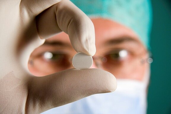 Eine behandschuhte Hand hält eine weiße Tablette zwischen Daumen und Zeigefinger; im Hintergrund unscharf eine Person mit OP-Maske und Haube. Fokus auf das Medikament.