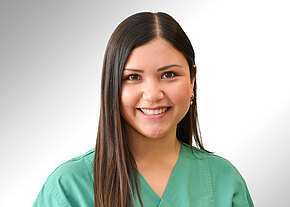 Porträt einer jungen Frau in grüner Pflege- bzw. OP-Kleidung, die freundlich in die Kamera lächelt. Neutraler, heller Hintergrund. Fokus auf ihr Gesicht und die medizinische Berufskleidung, suggeriert Gesundheitsberuf.