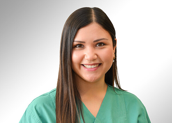 Porträt einer jungen Frau in grüner Pflege- bzw. OP-Kleidung, die freundlich in die Kamera lächelt. Neutraler, heller Hintergrund. Fokus auf ihr Gesicht und die medizinische Berufskleidung, suggeriert Gesundheitsberuf.