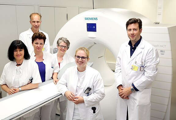 Sechs medizinische Fachkräfte in weißen Kitteln stehen vor einem Computertomografen. Sie lächeln in die Kamera, während sie sich in einer Klinik oder einem Krankenhaus befinden. Der Geräteschriftzug „SIEMENS“ ist sichtbar.