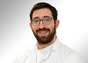 Porträt eines lächelnden Mannes mit Bart und Brille in weißem Kittel vor hellem Hintergrund, direkt in die Kamera blickend.