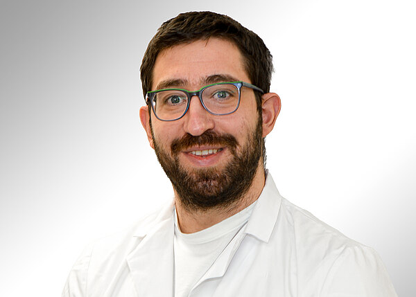 Porträt eines lächelnden Mannes mit Bart und Brille in weißem Kittel vor hellem Hintergrund, direkt in die Kamera blickend.