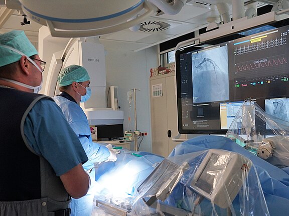 Zwei medizinische Fachkräfte führen im Herzkatheterlabor einen minimal-invasiven Eingriff durch. Sie verfolgen auf einem großen Monitor Röntgenbilder einer Koronararterie und EKG-Kurven, während der abgedeckte Patient auf dem Tisch liegt. Sterile Umgebung mit moderner Bildgebungstechnik.