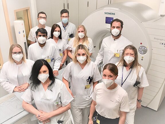 Ein Team von etwa zwölf medizinischen Fachkräften in weißer Arbeitskleidung und Masken steht in einem Radiologieraum vor einem großen Tomografen. Sie blicken in die Kamera als Gruppenfoto und vermitteln Professionalität und Zusammenarbeit.