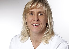 Station 5a Porträt einer lächelnden, mittelalten Frau mit schulterlangem blondem Haar in weißer Berufskleidung vor neutralem Hintergrund, frontal in die Kamera blickend.