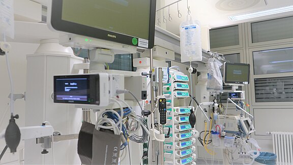 Helle Intensivstation mit Reihen moderner Medizingeräte: Monitore, Infusionspumpen, Beatmungsgerät, Schläuche und Kabel an Deckenschienen. Sterile, aufgeräumte Umgebung, bereit für intensivmedizinische Behandlung eines Patienten.