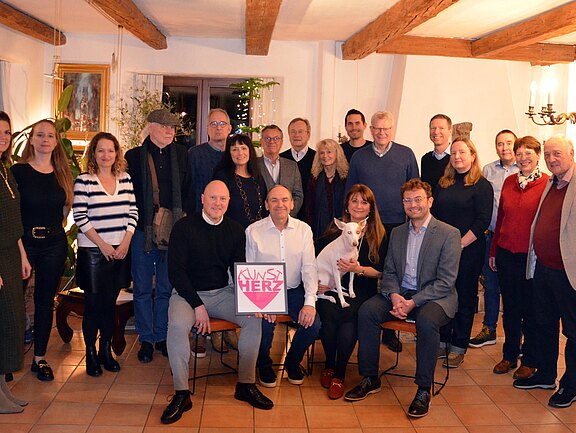 Gruppenfoto in einem hellen Raum: Mehrere Erwachsene posieren, einige sitzen vorn. Die Person in der Mitte hält ein Schild mit Herzsymbol und der Aufschrift „UNS HERZ“. Neben ihr sitzt jemand mit einem kleinen weißen Hund auf dem Schoß. Alle blicken lächelnd in die Kamera.