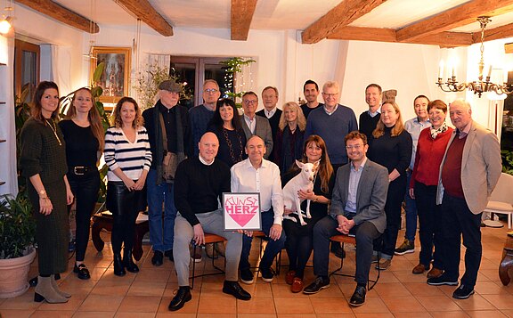 Gruppenfoto in einem hellen Raum: Mehrere Erwachsene posieren, einige sitzen vorn. Die Person in der Mitte hält ein Schild mit Herzsymbol und der Aufschrift „UNS HERZ“. Neben ihr sitzt jemand mit einem kleinen weißen Hund auf dem Schoß. Alle blicken lächelnd in die Kamera.