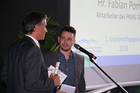 Zwei Männer auf einer Bühne während einer Preisverleihung: Einer überreicht dem anderen Geschenk und Umschlag am Mikrofon. Im Hintergrund zeigt die Leinwand eine Präsentationsfolie mit der Aufschrift „Krankenpflegepreis 2018“.