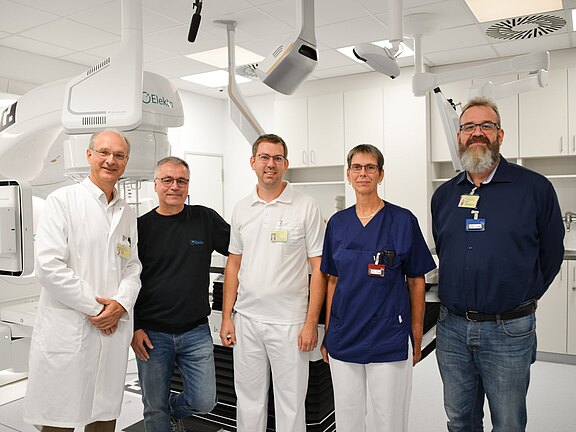 Fünf Personen stehen in einem modernen medizinischen Raum, der mit komplexen Geräten ausgestattet ist. Sie tragen verschiedene Berufskleidung: ein Arzt in einem weißen Kittel, Techniker in einem schwarzen T-Shirt, ein weiterer Arzt in weißer Uniform und zwei Personen in blauen und schwarzen Hemden.