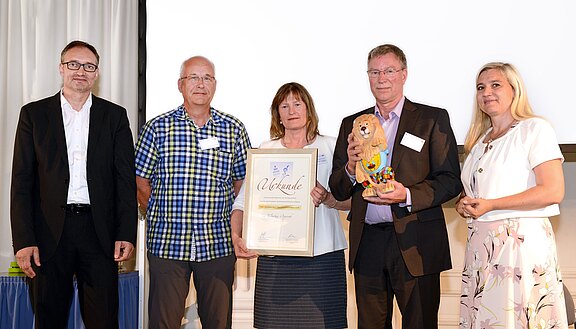 Fünf Erwachsene posieren bei einer Preisverleihung; die Frau in der Mitte hält eine gerahmte Urkunde, der Mann daneben eine bunte Bärenfigur als Trophäe. Alle stehen vor hellem Hintergrund und blicken in die Kamera.