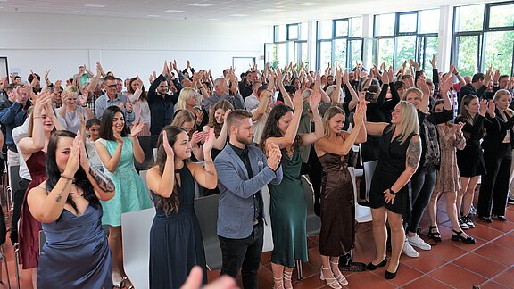 Eine große Gruppe von Menschen steht in einem Raum und applaudiert begeistert. Die Anwesenden tragen verschiedene formelle Kleidung und zeigen Freude. Die Atmosphäre ist festlich und es scheint ein besonderes Ereignis zu feiern. Tageslicht strömt durch große Fenster.