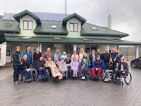 Großes Gruppenfoto vor einem Gebäude: Menschen unterschiedlichen Alters, viele in Rollstühlen, einige stehend. Sie lächeln und posieren zusammen, als Teil einer inklusiven Gemeinschaft oder Veranstaltung.