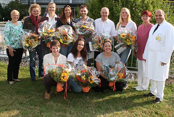 Gruppenfoto im Freien: Zwölf Erwachsene, einige tragen Krankenhauskleidung. Vordere Reihe kniet, alle halten große Blumensträuße und lächeln. Feierlicher Anlass, vermutlich Ehrung von Mitarbeitenden.
