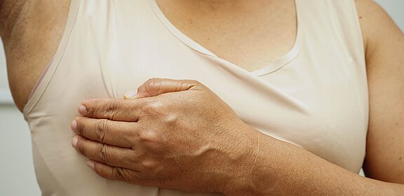 Eine Person in ärmellosem Oberteil hebt den Arm und tastet mit der anderen Hand die Brust ab – eine Selbstuntersuchung zur Kontrolle auf Knoten bzw. Brustkrebs-Anzeichen.