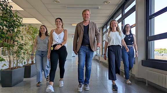 Fünf junge Erwachsene gehen nebeneinander lächelnd auf die Kamera zu. Heller Büroflur mit großen Fenstern. Lässige Kleidung, entspannte Stimmung; wirkt wie ein Team auf dem Weg zu einem Termin.