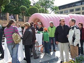 Eine Gruppe von Menschen steht um Stehtische und unterhält sich. Im Hintergrund befindet sich eine große, rosa Struktur. Die Szene spielt sich im Freien ab, umgeben von Bäumen und Gebäuden. Die Atmosphäre wirkt gesellig und freundlich.