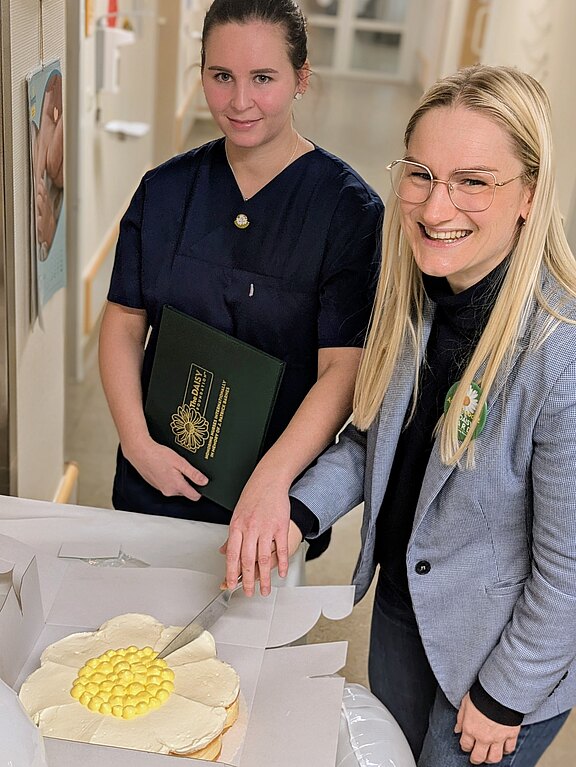 Zwei Frauen in einem Flur: Eine in dunkelblauer Arbeitskleidung hält eine Mappe, die andere mit Brille lächelt und schneidet eine große, blumenförmig dekorierte Torte an. Feierlicher Moment.