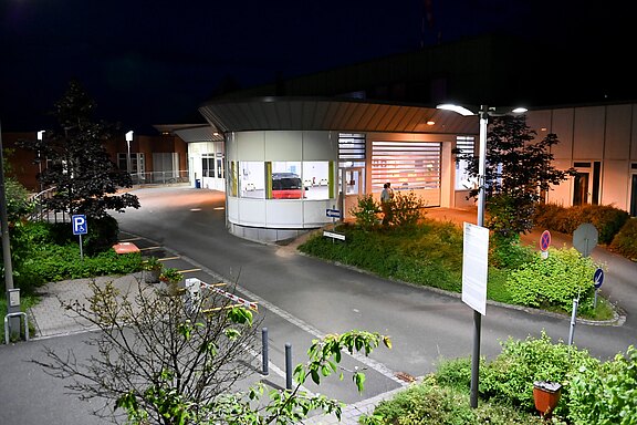 Nachtszene: Hell erleuchteter Eingangsbereich eines Krankenhauses mit runder Vorfahrt und breiter Schiebetür. Leere Zufahrt, wenige Wegweiser und Straßenlampen, umgeben von Sträuchern.