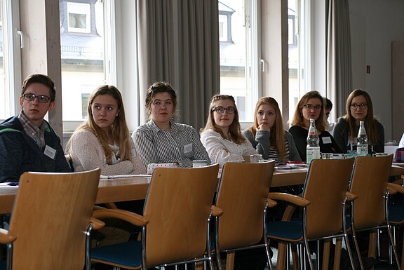 Gruppe junger Erwachsener sitzt an einem langen Konferenztisch und blickt aufmerksam nach links, als hörten sie einem Vortrag zu. Vor ihnen liegen Notizen und Getränke.