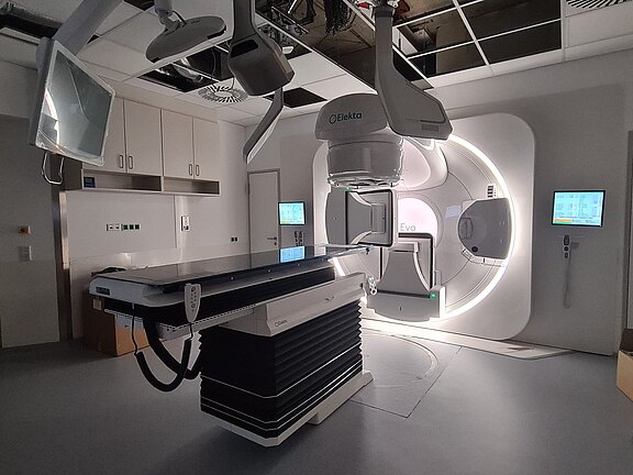 Beleuchteter Behandlungsraum mit modernem Linearbeschleuniger für Strahlentherapie: ringförmiger Strahlkopf, drehbarer Gantry, höhenverstellbare Patientenliege und Kontrollmonitore in klinischer Umgebung.