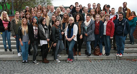 Großes Gruppenfoto junger Erwachsener auf Außentreppen. Mehrere Reihen, überwiegend junge Frauen, einige Männer, in Freizeitkleidung. Alle blicken zur Kamera und lächeln. Wirkt wie eine Schul- oder Studiengruppe bei einem Treffen im Freien.