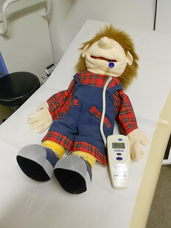 Eine große Handpuppe in Latzhose liegt auf einer Untersuchungsliege. Ein medizinischer Sensor mit Kabel steckt am Mund; der tragbare Monitor liegt daneben. Darstellung einer kindgerechten Untersuchung oder Demonstration.