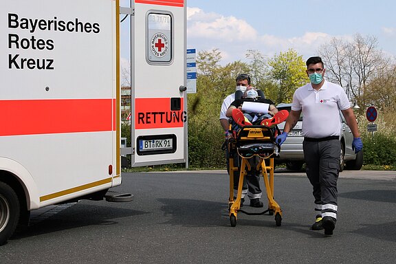 Ein Rettungssanitäter trägt eine Trage mit einer Person, während ein weiterer Sanitäter assistiert. Im Hintergrund ist ein Fahrzeug des Bayerischen Roten Kreuzes zu sehen. Beide Sanitäter tragen Schutzmasken und Handschuhe.