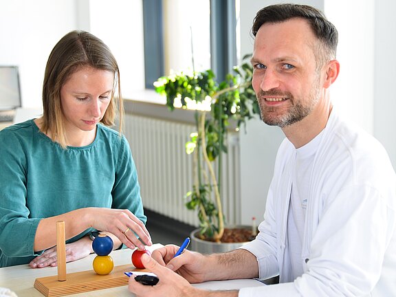 Zwei Personen bei einer Ergotherapie-Übung. Eine Frau stapelt farbige Holzkugeln auf einen Holzstab, der Mann daneben lächelt und schreibt Notizen. Helles Praxisumfeld.