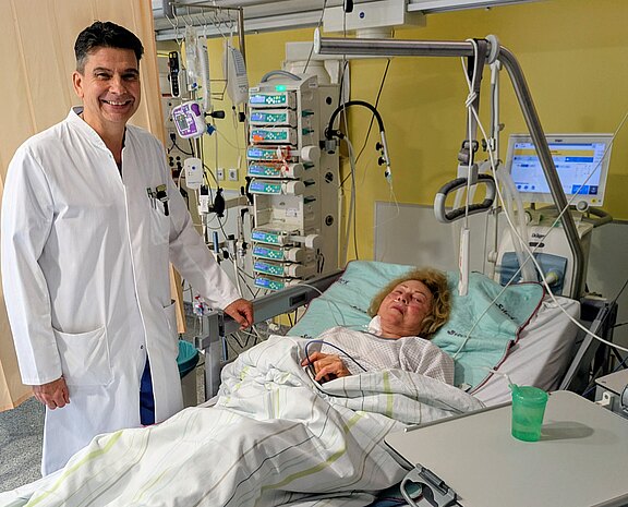 Arzt in weißem Kittel steht lächelnd neben einer Patientin im Krankenhausbett. Die Frau liegt zugedeckt, an Überwachungsgeräte und Infusionen angeschlossen. Krankenhausmonitor und Pumpen zeigen medizinische Werte. Szene vermittelt klinische Betreuung und Überwachung auf einer Intensivstation oder Überwachungsstation.