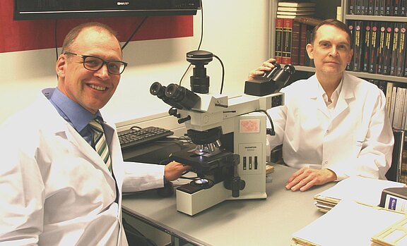 Zwei Forschende in weißen Kitteln sitzen im Labor an einem großen Lichtmikroskop. Eine Person lächelt zur Kamera, die andere hält das Okular. Schwerpunkt: gemeinsame mikroskopische Forschung.